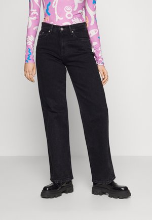 Osoba ubrana w czarne, wysokowcieniowe szerokie jeansy, fioletową wzorzystą bluzkę z długim rękawem oraz czarne masywne buty na platformie, stojąca na szarej podłodze.