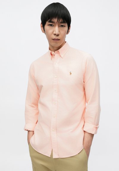 Ανοιχτό ροζ πουκάμισο με κουμπιά, κατασκευασμένο από ύφασμα με απαλή υφή. Διαθέτει μικρό κεντητό λογότυπο στο αριστερό στήθος και μανίκια που είναι σηκωμένα.