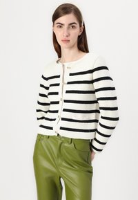 Strikket cardigan med hvit base og sterke svarte striper; har rund hals, gullknapper og to frontlommer. Kombinert med grønne bukser.