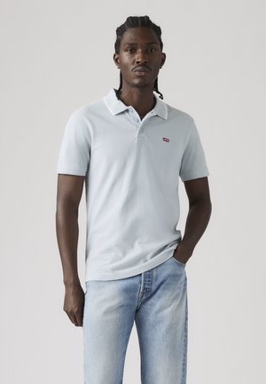 Hombre con trenzas, vestido con una camiseta polo azul claro de Levi’s y vaqueros azules desteñidos, de pie con una mano en el bolsillo frente a un fondo liso.