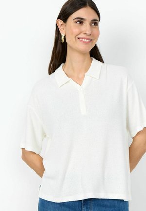 Žena s dugom smeđom kosom koja nosi zlatne naušnice u obliku prstena i bijelu pletenu polo majicu kratkih rukava, smiješi se i gleda desno.