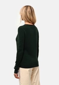 Maglione verde scuro con scollo rotondo e maniche lunghe, realizzato in materiale morbido. Presenta polsini aderenti e una trama liscia. Abbinato a pantaloni chiari.