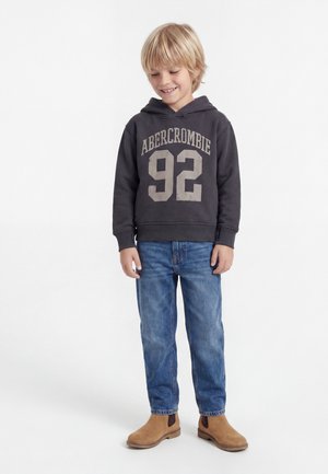 Smilende ung dreng iført en mørk hættetrøje med "Abercrombie 92," blå jeans og brune støvler, stående foran en hvid baggrund.