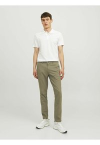 Witte polo met korte mouwen, gecombineerd met olijfgroene chino's. Schoenen zijn witte sneakers. Effen achtergrond.