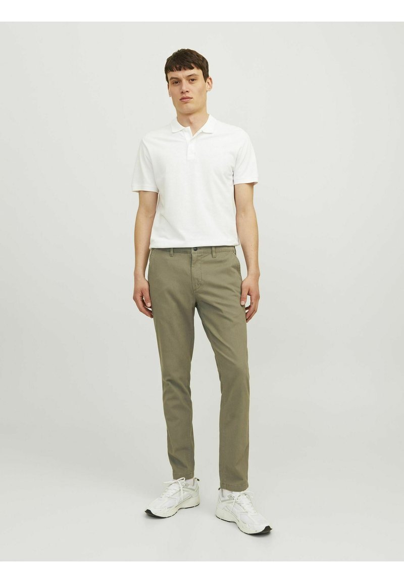Camisa polo blanca de manga corta combinada con pantalones chinos verde oliva. El calzado son zapatillas blancas. Fondo plano.