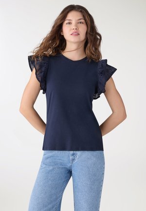 Giovane donna con capelli castano ondulati che indossa un top senza maniche navy con spalle in pizzo arricciato e jeans azzurri, in piedi contro uno sfondo semplice.