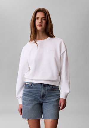 Młoda kobieta z długimi brązowymi włosami, ubrana w białą bluzę Calvin Klein i niebieskie dżinsowe szorty na jednolitym szarym tle.