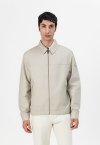Beige lichtgewicht jack met een rits aan de voorkant, kraag, zijzakken en elastische manchetten; heeft een subtiele horizontale naad over de borst.
