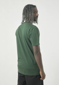 Grünes kurzärmeliges T-Shirt aus weichem Stoff, mit Rundhalsausschnitt; Modell zeigt nach links mit texturiertem Haar und schwarzen Hosen.