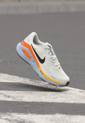 Hvid Nike løbesko med sort swoosh, orange og blå sål, svævende over asfalt med små stykker affald nedenunder.