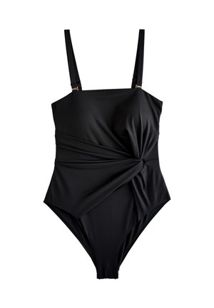 Žena kučeravých vlasov v čiernych jednodielnych plavkách stojí vedľa zakrivenej bielej textúrovanej steny pod jasnou modrou oblohou.