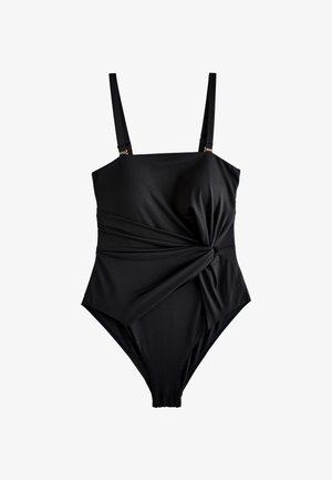 Žena s kovrčavom kosom u crnom jednodijelnom kupaćem kostimu stoji pored zakrivljenog bijelog teksturiranog zida pod vedrim plavim nebom.