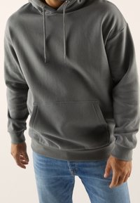 Felpa grigia con cappuccio realizzata in tessuto morbido, con tasca frontale, lacci e polsini a costine. Indossata con jeans blu.