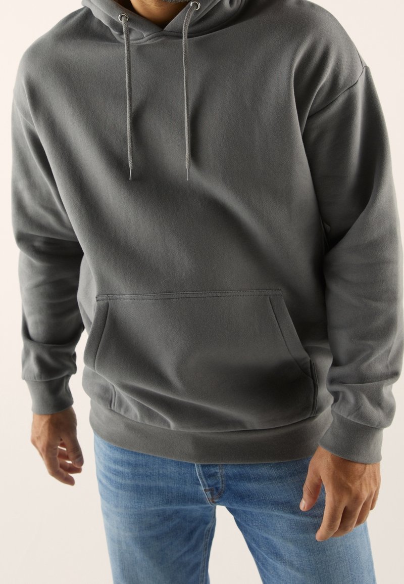 Felpa grigia con cappuccio realizzata in tessuto morbido, con tasca frontale, lacci e polsini a costine. Indossata con jeans blu.