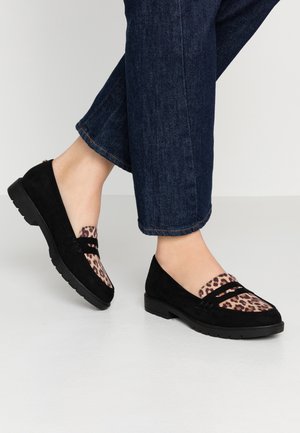 Pieds portant des mocassins noirs avec un imprimé léopard sur le dessus, associés à un jean en denim bleu foncé.