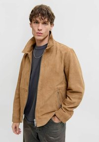 Tan ruskindsjakke med ståkrave, frontlynlås og sidelommer med lynlås. Bæres over en mørkegrå skjorte. Glat tekstur, afslappet design.