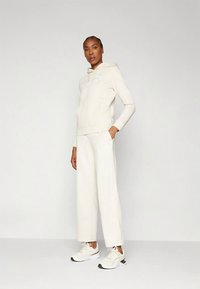 Bēša krāsas sporta džemperis ar kengūras kabatu, atbilstošas plaša silueta bikses un neitrālas krāsas sporta apavi. Priekšpusē ir logotipa apdruka.