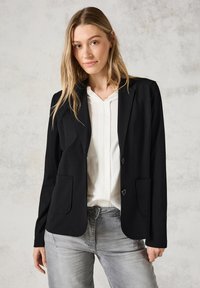 Blazer nero con chiusura a un bottone, due tasche frontali e maniche lunghe. Indossato sopra una camicia bianca, con una texture morbida e liscia.