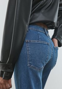 Korkeavyötäröiset siniset farkut, joissa on sileä tekstuuri, klassinen viiden taskun muotoilu ja kontrastiväriset tikkaukset. Yhdistetty mustaan nahkapaitaan.