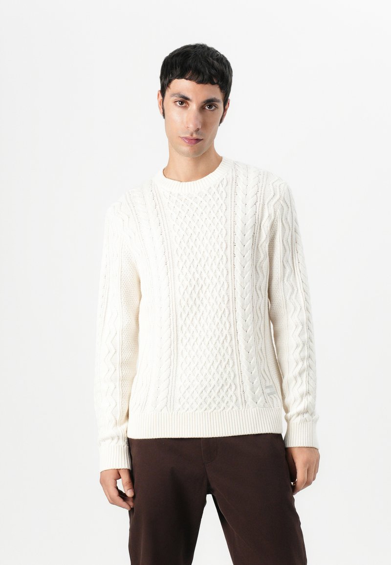 Kabelstrikket creme sweater med ribbede manchetter og kant. Har strukturerede mønstre og en klassisk rund hals. Sammen med mørke bukser.