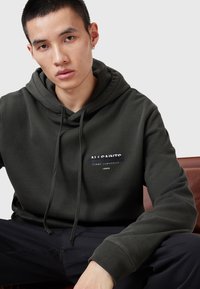 Donkergroene hoodie met een kangoeroezak, trekkoordkap en logo-detail aan de voorkant in het wit. Zacht materiaal, relaxte pasvorm, lange mouwen.