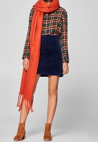 Chemise à carreaux verte et orange, jupe en velours côtelé bleu marine, écharpe orange vif avec franges, et bottines brunes.