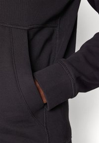 Main portant une manche de sweat-shirt foncé placée dans la poche kangourou avant avec des détails de couture visibles.