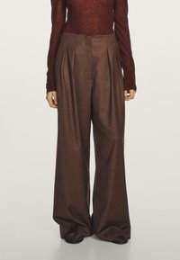 Brązowe spodnie o szerokich nogawkach wykonane z teksturowanej tkaniny, z plisami i wysokim stanem. Brak widocznych kieszeni lub ozdób.