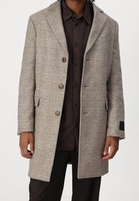 Ternet frakke i beige toner lavet af tekstureret stof med en krave med hakket slag, tre knapper og sidelommer. Mørk skjorte synlig under.