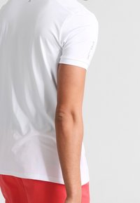 Bílý přiléhavý tričko s krátkým rukávem, vyrobené z lehkého materiálu, s jemným logem na rameni.
