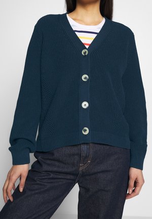 Vrouw draagt een donkerblauwe geribde gebreide cardigan met vier doorschijnende knopen, gecombineerd met donkere denim jeans en een gestreept overhemd eronder.