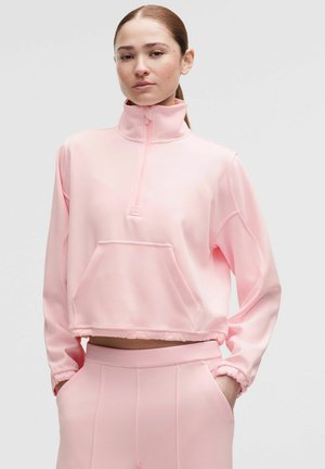 Młoda kobieta ubrana w jasno różowy krótki sweter z półzamkiem i pasujące wysokie spodnie z wysokim stanem, ręce w kieszeniach, neutralne tło.