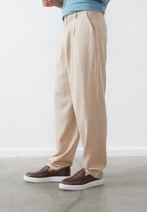 Beige, weit geschnittene Hose mit Falten, aus strukturiertem Stoff. Kombiniert mit braunen Slippers mit weißen Sohlen.