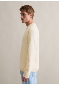 Cremefarbener strukturierter Pullover mit rundem Kragen und gerippten Bündchen, kombiniert mit hellblauen Jeans, von der Seite gezeigt.