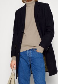 Schwarzer Wollmantel mit tailliertem Design, drapiert über einem beigen Strickpullover mit Rollkragen. Blaue Jeans und eine beigefarbene Tragetasche sind sichtbar.