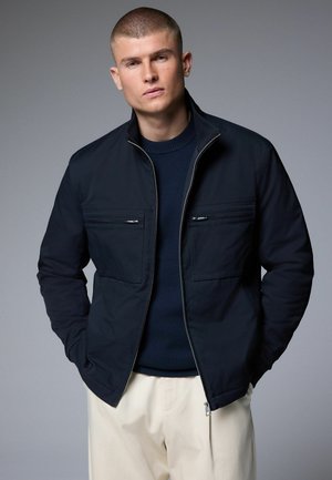 Navy zip-up jacket with two front pockets and a high collar, paired with a dark sweater and light trousers. Smooth fabric texture.