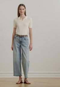 Adīta krēmkrāsa polo krekls ar ribota tekstūru, saskaņots ar gaišziliem platām biksēm un brūnām sandalēm ar minimālistisku dizainu.