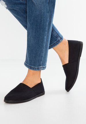 Schwarze Canvas-Slipper mit niedrigem Profil, runder Zehenpartie und flexiblen Gummisohlen, präsentiert zusammen mit Jeans.