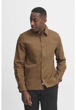 Brown cotton button-up shirt with two chest pockets, long sleeves, and a collared neck, shown on a person against a plain background.