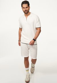 Kurzärmliges weißes Polohemd mit Reißverschluss und schwarzem Besatz, kombiniert mit weißen strukturierten Shorts und beigen Sneakers; Modell steht vor einem neutralen Hintergrund.
