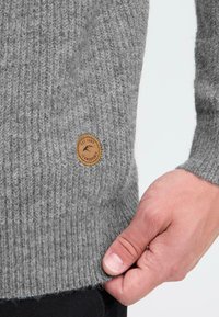 Grijze geribbelde gebreide trui met een ronde, bruine leren patch met het label "est. 1991" aan de onderkant. De textuur lijkt zacht en dik te zijn.