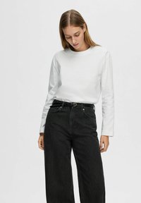 Camicia bianca a maniche lunghe abbinata a jeans neri a vita alta e a gamba larga. La camicia presenta un collo rotondo e una consistenza liscia.