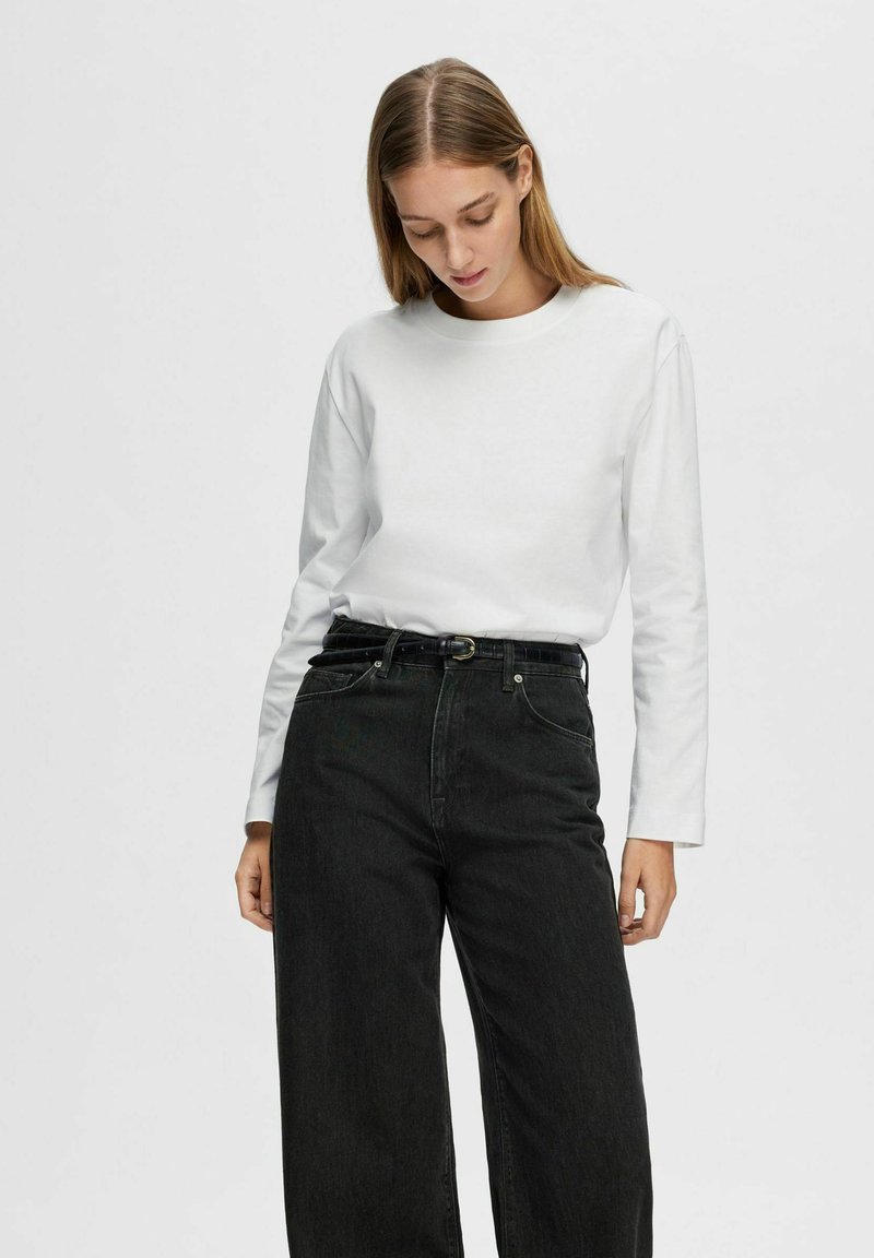 Camicia bianca a maniche lunghe abbinata a jeans neri a vita alta e a gamba larga. La camicia presenta un collo rotondo e una consistenza liscia.