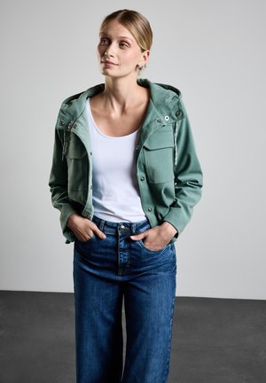 Giacca in cotone verde con cappuccio e due tasche frontali, abbinata a una maglietta bianca e a jeans a gamba larga in denim blu. Tessuto texture e dettagli con bottoni.