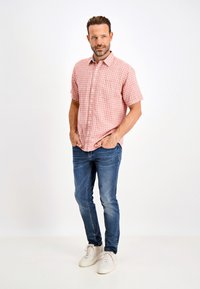 Camisa de botonada en patrón de cuadros rosa claro, de mangas cortas. Llevada con jeans de lavado medio y zapatillas blancas, con las manos en los bolsillos.