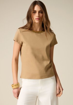 Kvinde med langt brunt hår iført en tan kortærmet top med guldpaliet-halsudskæring, hvide bukser og to gyldne armbånd på venstre håndled.