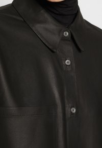 Chemise en cuir noire avec col, devant boutonné, poche visible, portée sur un col roulé noir.