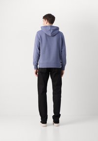 Felpa con cappuccio azzurro chiaro, dotata di polsini e orlo a coste, abbinata a pantaloni neri e scarpe da ginnastica. La felpa presenta una vestibilità comoda e una vista posteriore.