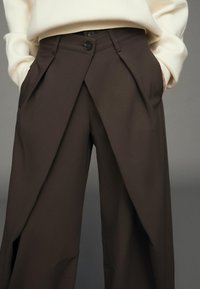 Hnědé, na míru šité kalhoty s předním skladem a zapínáním na jeden knoflík. Mají boční kapsy a hladkou, strukturovanou látku.