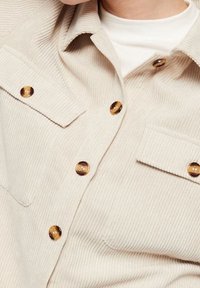 Beige kordflöjtsjacka med sköldpaddsmönstrade knappar och fickor med lock på bröstet, worn över en vit t-shirt med rund halsringning.